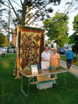 Photo of 2 ladies standing by a table set up in front or a large hanging quilt, taken at a Thursday Evening Summer Band Concert on the Common Thank You to Ed West